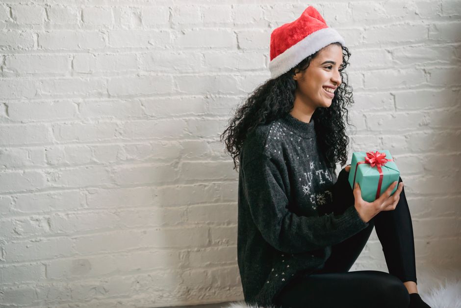 Side view content ethnic female in soft sweater and red Santa hat sitting on floor with wrapped gift box and looking away with happy smile during Christmas celebration