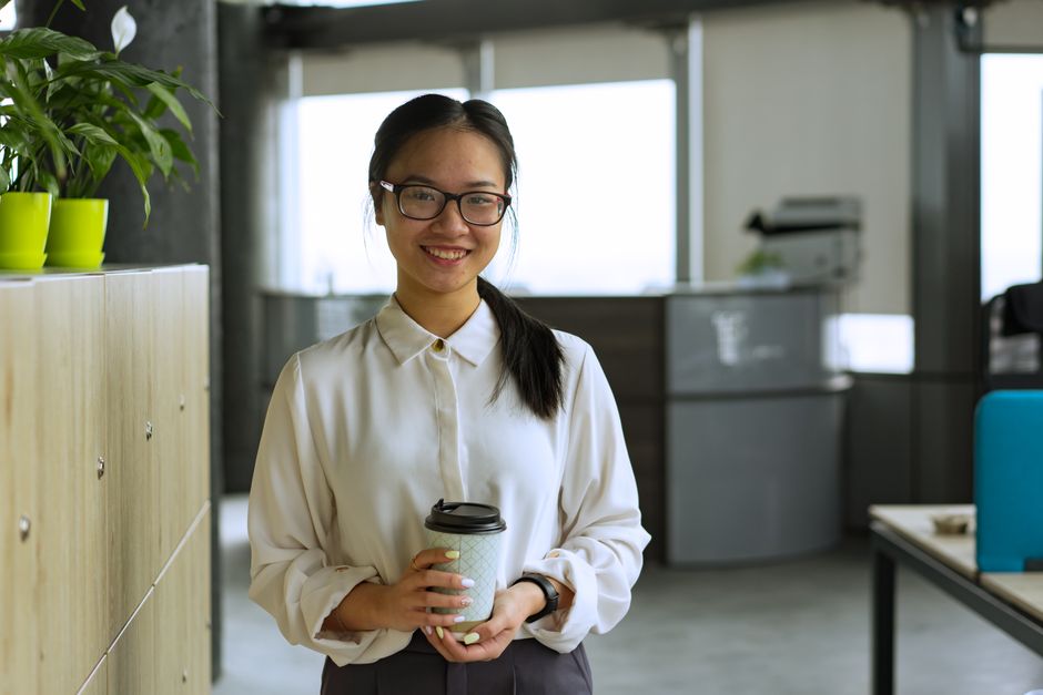 Smiling woman with glasses holding a coffee cup in a modern office setting.