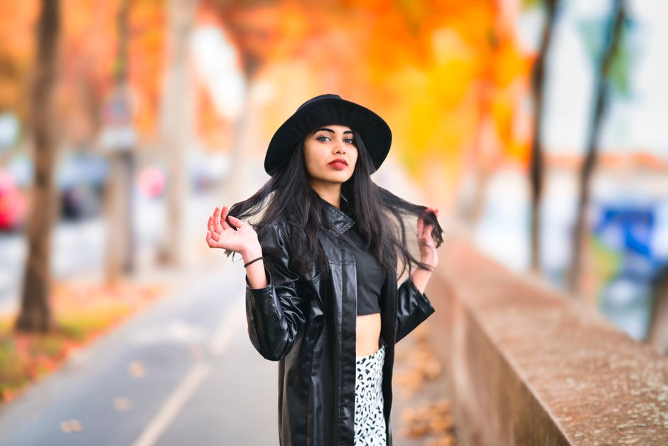 A fashionable woman in a black hat poses confidently outdoors with vibrant autumn foliage in the background