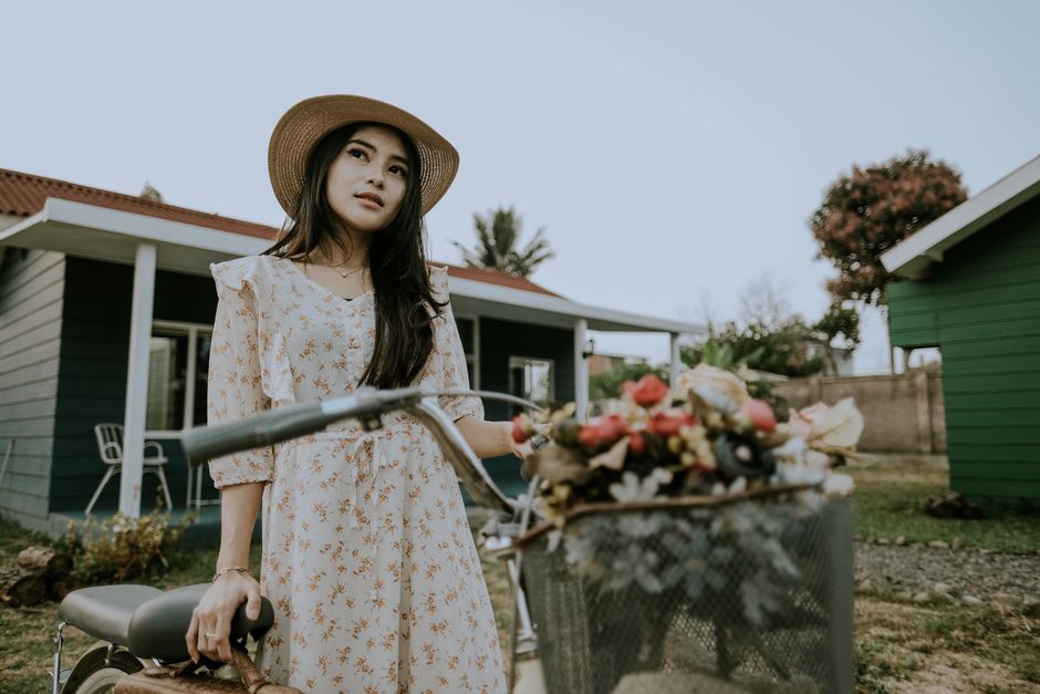 A stylish woman in a summer dress stands with a bicycle in a garden setting