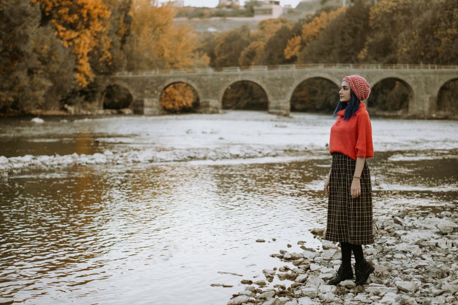 A woman stands on a rocky shore by a river with a stone bridge, surrounded by fall foliage