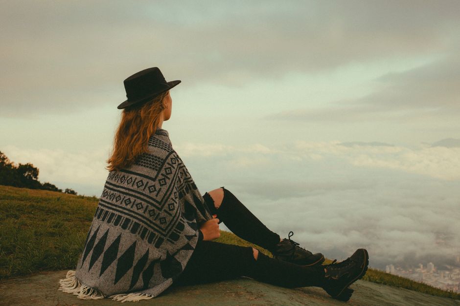 A young woman in a patterned poncho and hat sits on a hill, enjoying a panoramic view in Jaraguá do Sul, Brazi