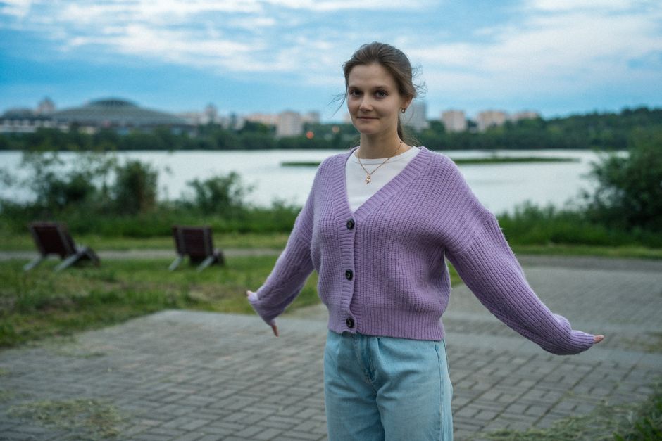 A young woman in a purple sweater poses by a lake with a cityscape backdrop