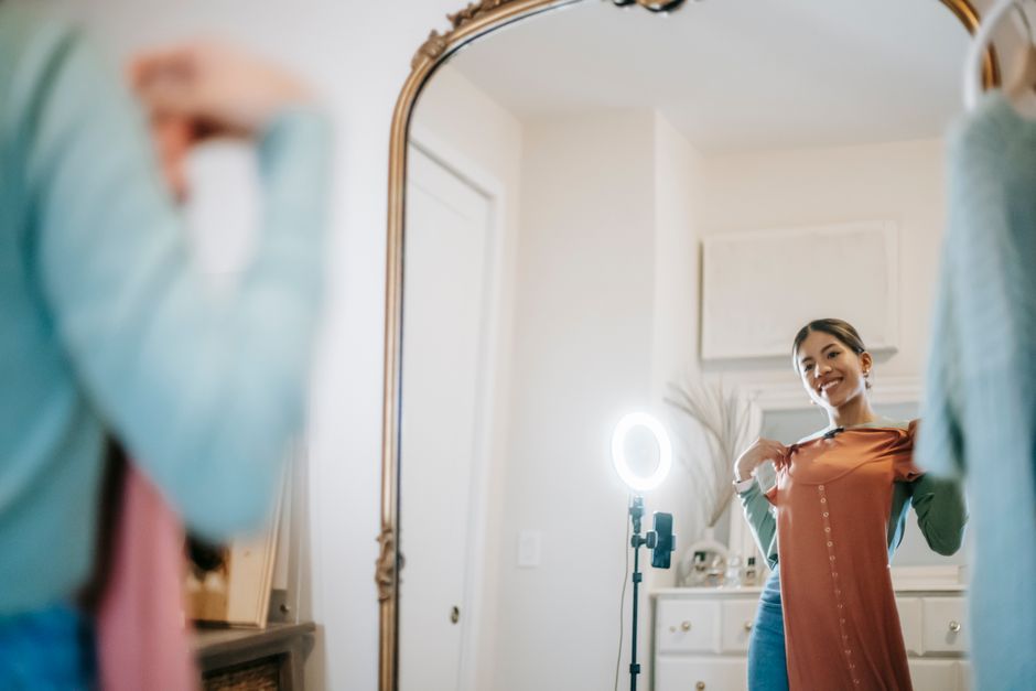 Positive young ethnic female blogger in trendy outfit standing in room and demonstrating pink dress while looking at mirror and shooting video on phone with ring lamp near cabinet