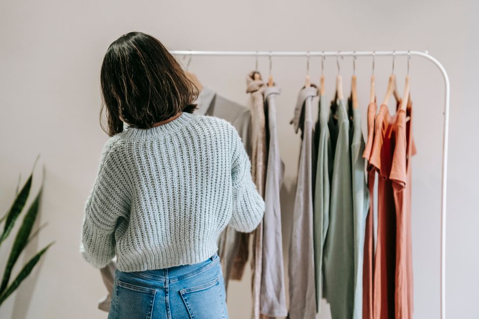 Back view of anonymous female in knitted sweater against stand with collection of wear on light background