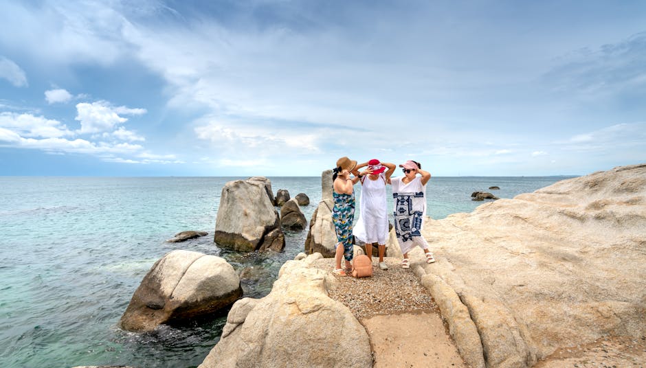 Three women in summer attire standing by the rocky seashore under an overcast sky.