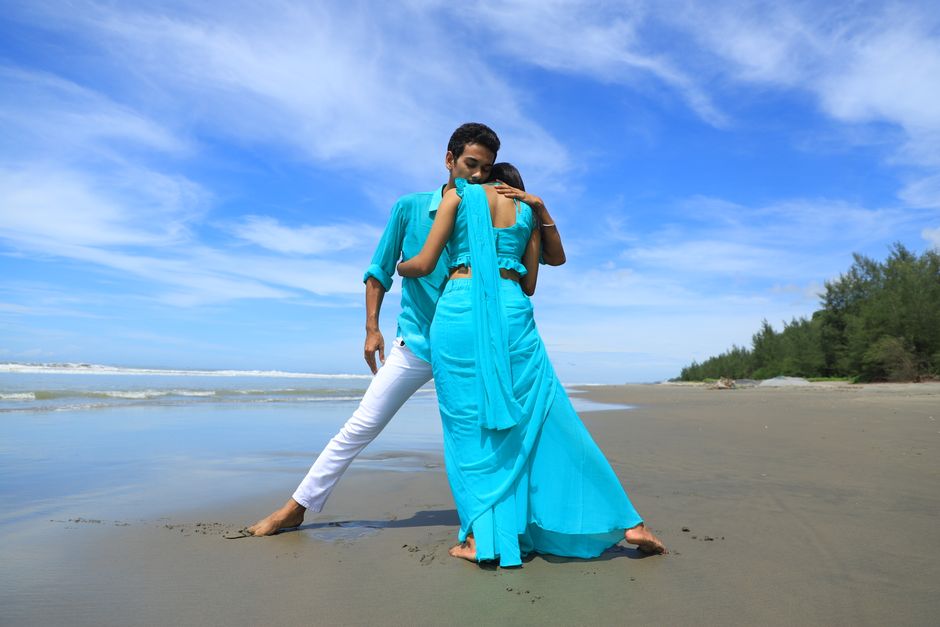 Young couple in turquoise outfits embracing on a serene beach under a bright blue sky.