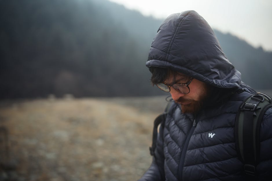 A man in a hooded jacket, lost in thought, stands amidst a misty mountain backdrop.