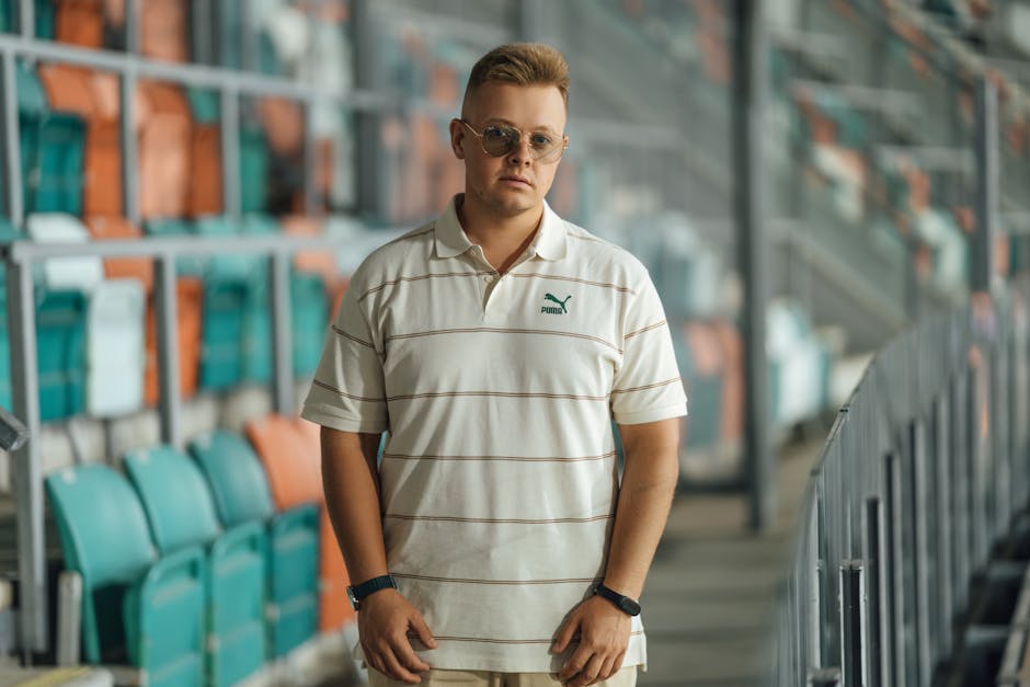 A young man with sunglasses in a casual outfit stands in a deserted sports stadium.