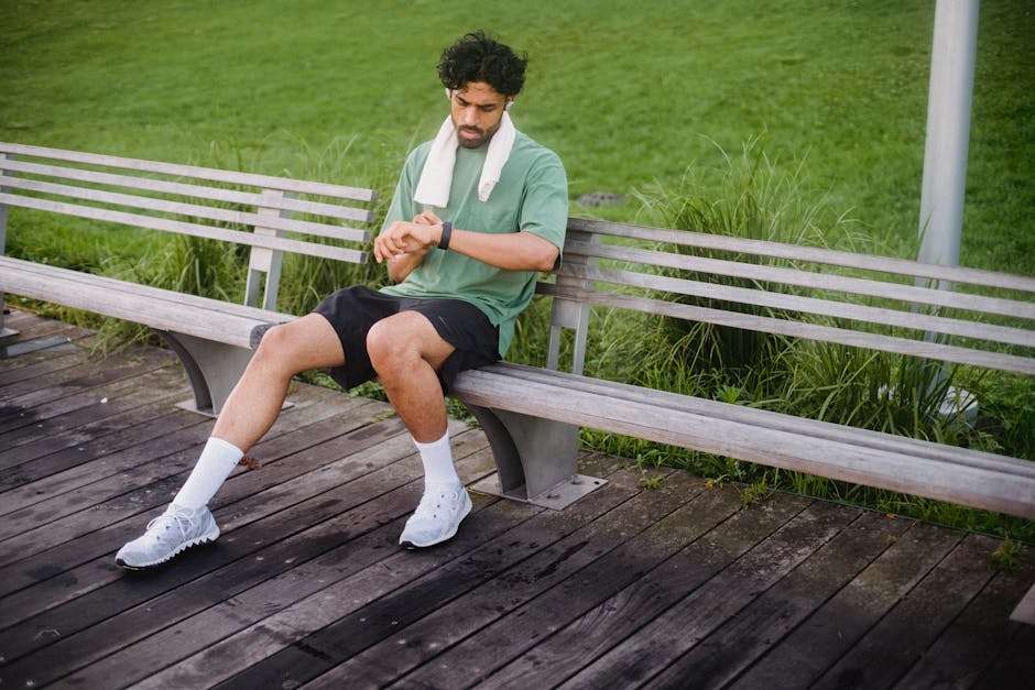 A man in activewear rests on a bench while checking his fitness tracker in a park.