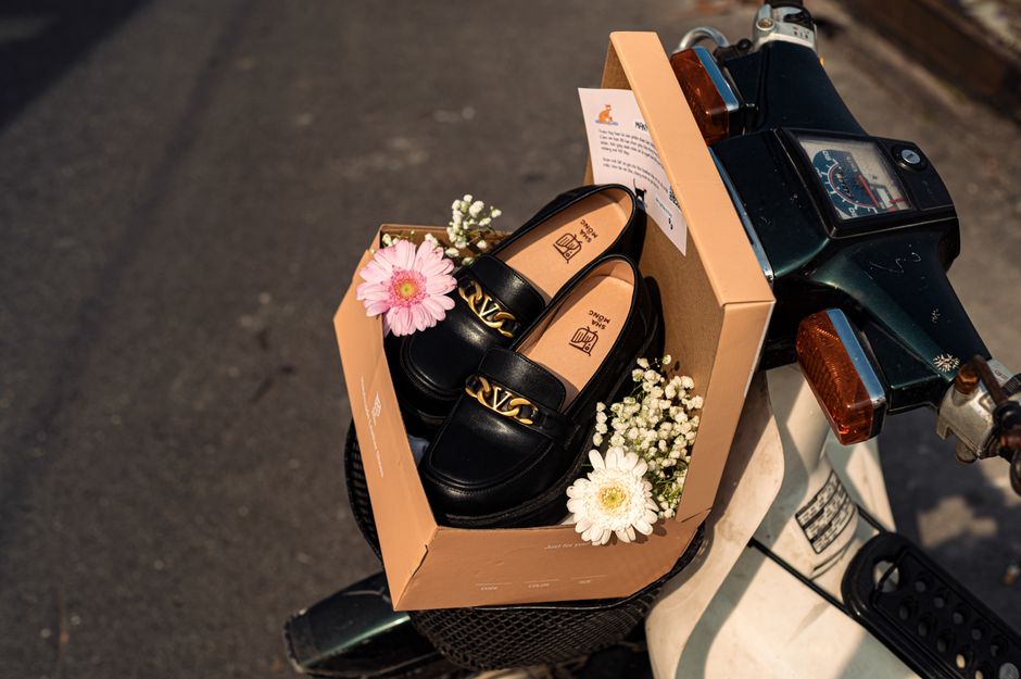 Black loafers in a brown box adorned with flowers, placed on a vintage scooter