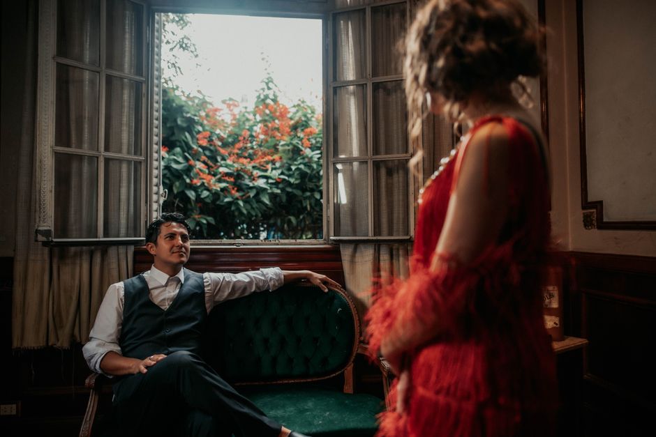 A man seated in a vintage room, gazing at a woman in a red dress by an open window.