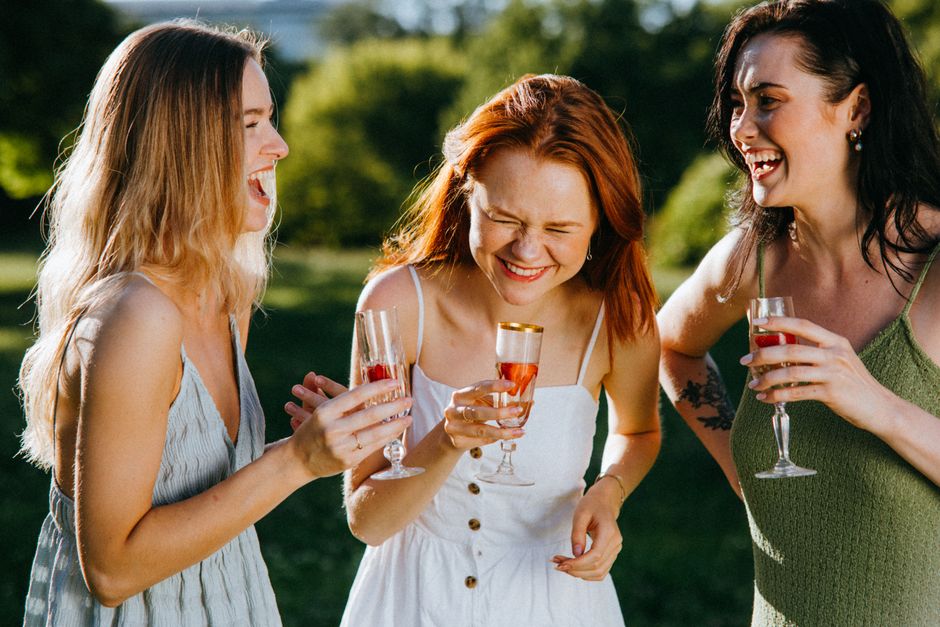 Three women laugh and enjoy wine outdoors, celebrating friendship on a sunny day.