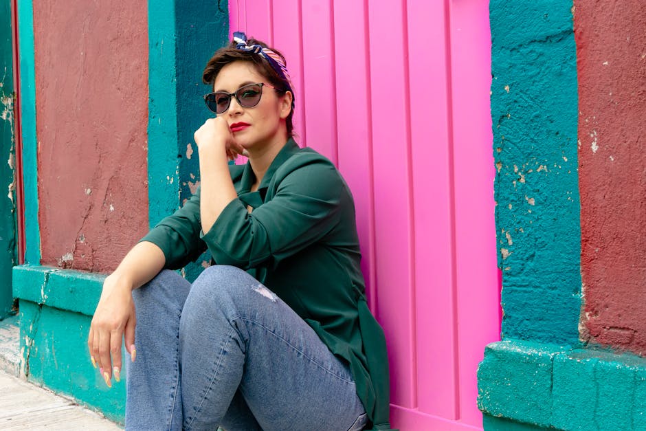 Chic woman in denim and green blazer sitting by a vibrant pink wall, wearing sunglasses and a kerchief.