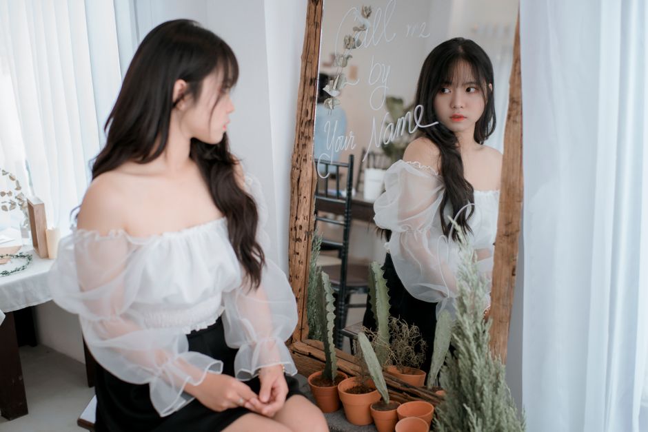 Elegant woman in white blouse looking in mirror indoors