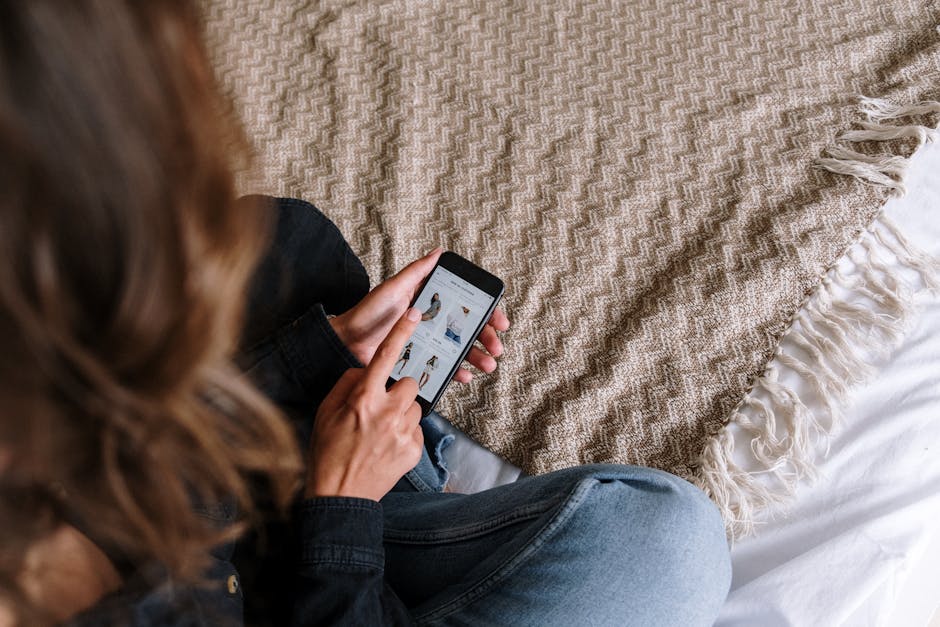 A woman browsing online shopping apps on her smartphone while sitting on a bed with a cozy blanket.