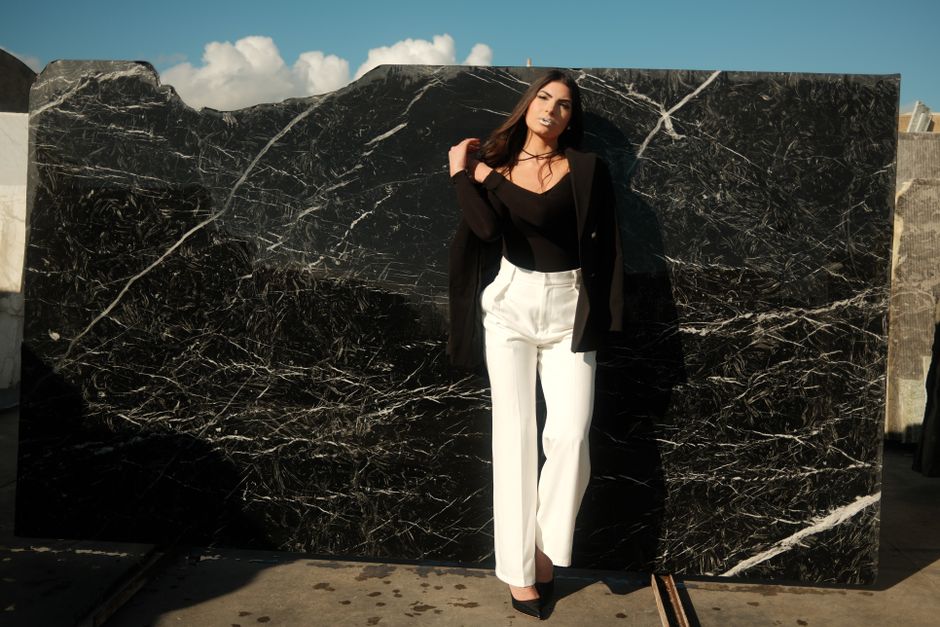 Fashionable woman in black top and white pants posing in sunlight against a striking black marble background