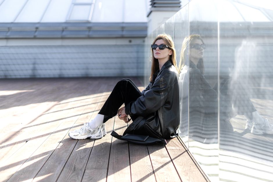 Fashionable woman in sunglasses and black outfit sits against a glass wall with urban surroundings