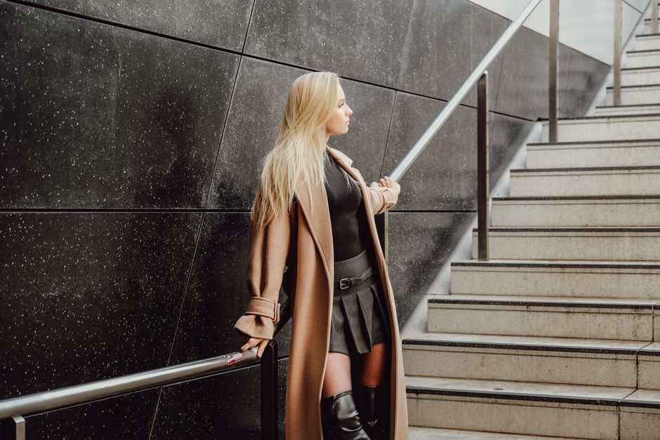Fashionable woman in trench coat and leather boots standing by a modern staircase