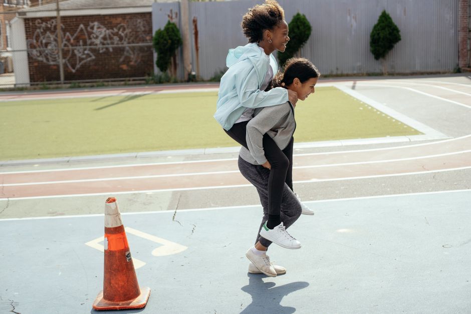 Side view of cheerful diverse teenage girls doing piggyback ride on sports ground during physical education class