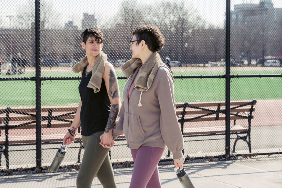 Girlfriends in sport outfits passing by sports ground while looking at each other and holding hands in daylight