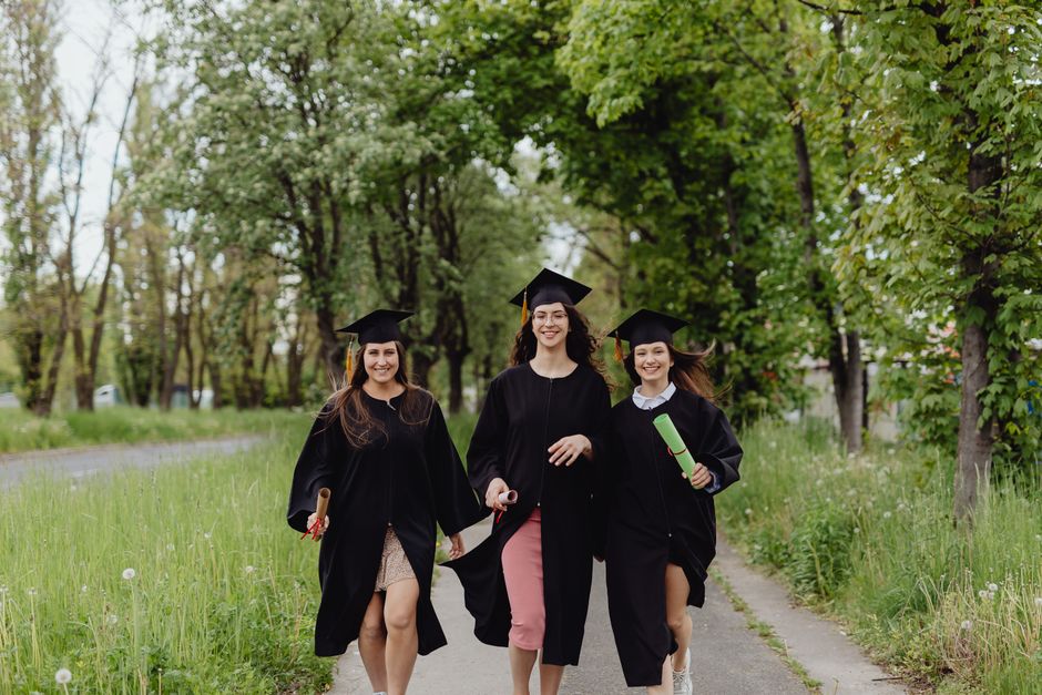 Three female graduates in academic robes, celebrating outdoors in a lush green environment.