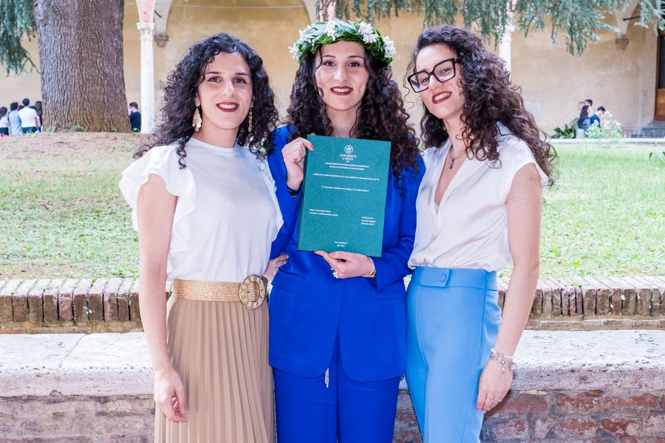 Three women posing together outdoors, one holding a graduation diploma.