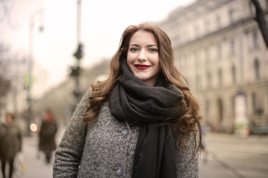 Happy woman in a gray coat and scarf standing outdoors, showcasing a charming smile on a city street