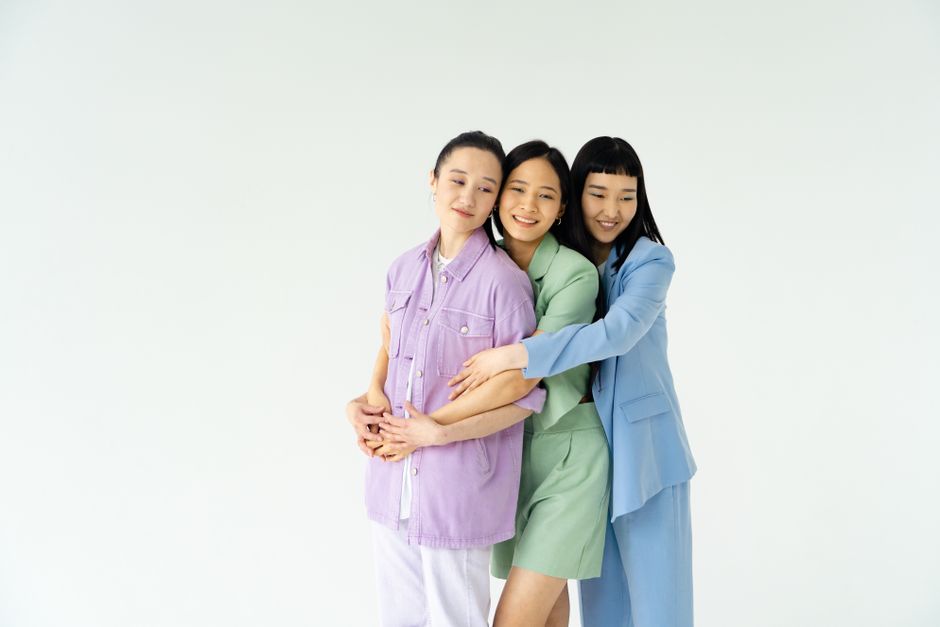 Three women in pastel outfits embrace against a white background, symbolizing friendship and affection.