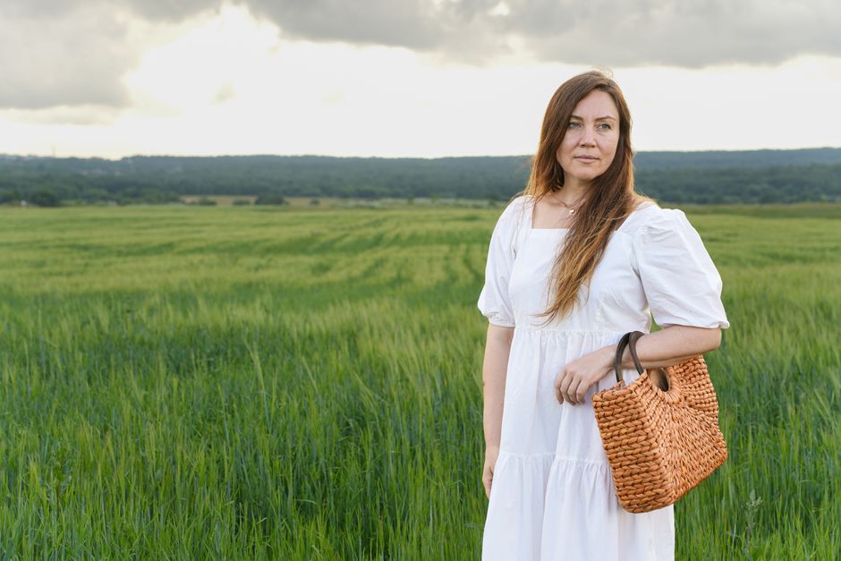 A woman in a white dress stands in a green field, holding a woven wicker bag under a cloudy sky.