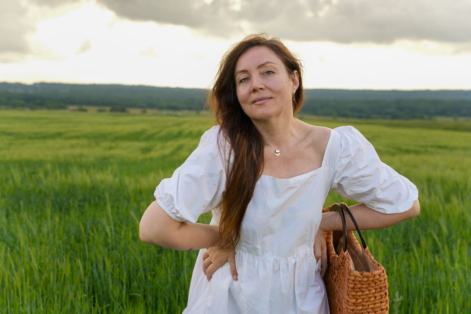A woman in a white dress stands relaxed in a lush green field, exuding tranquility.