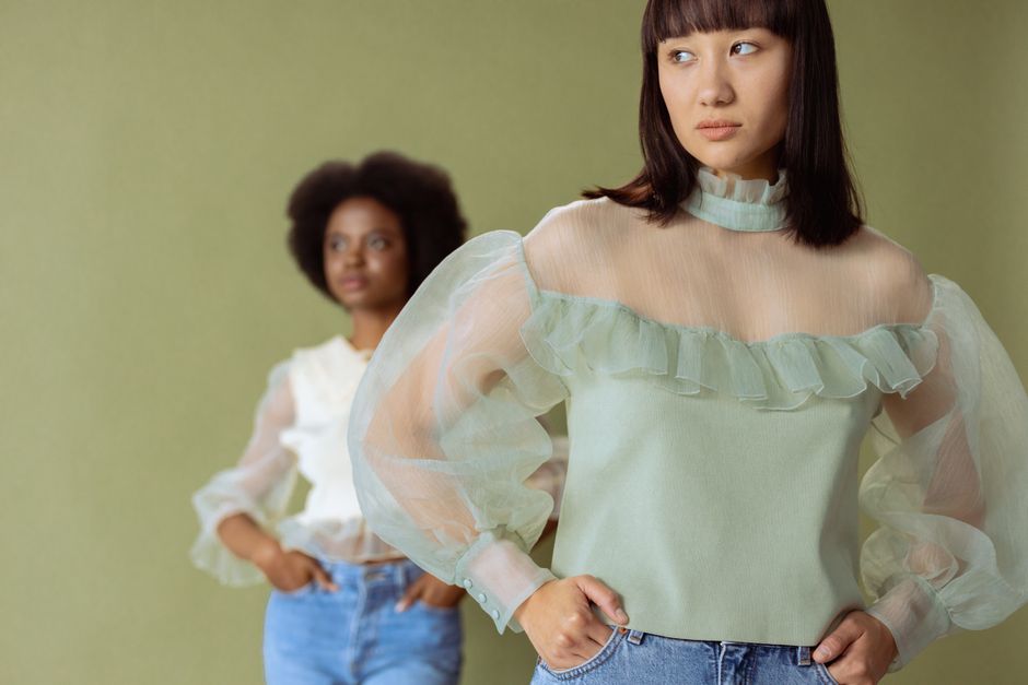 Two women in sheer blouses pose in a stylish studio shot with a subtle green background