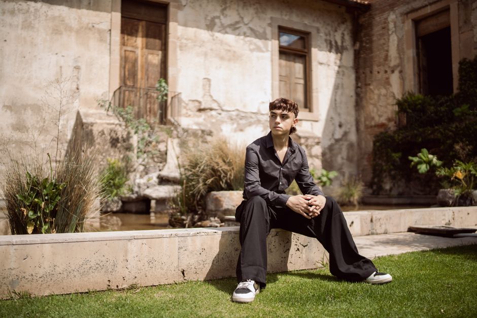 Young man in black shirt sitting outdoors by a rustic building, showcasing fashion style.