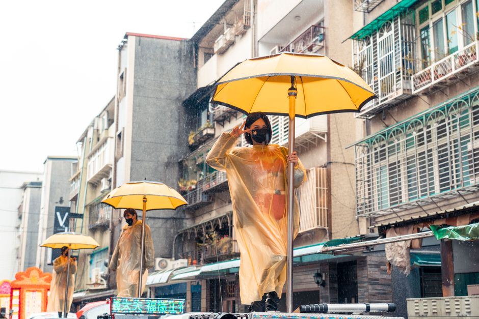 Women in raincoats and face masks hold yellow umbrellas during a rainy day parade in an urban setting