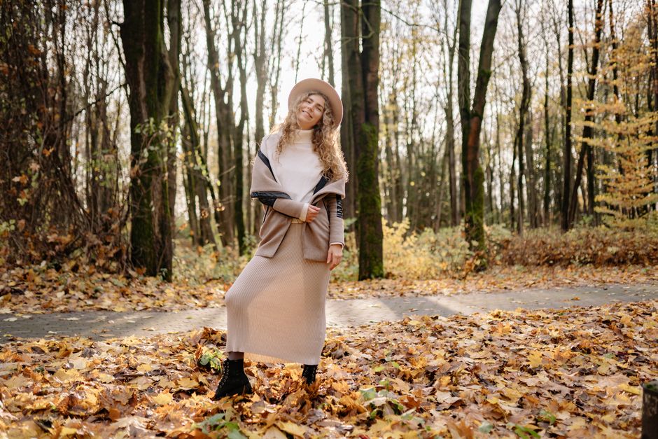 Young woman with curly hair smiling joyfully in an autumn park filled with colorful leaves, wearing a stylish 